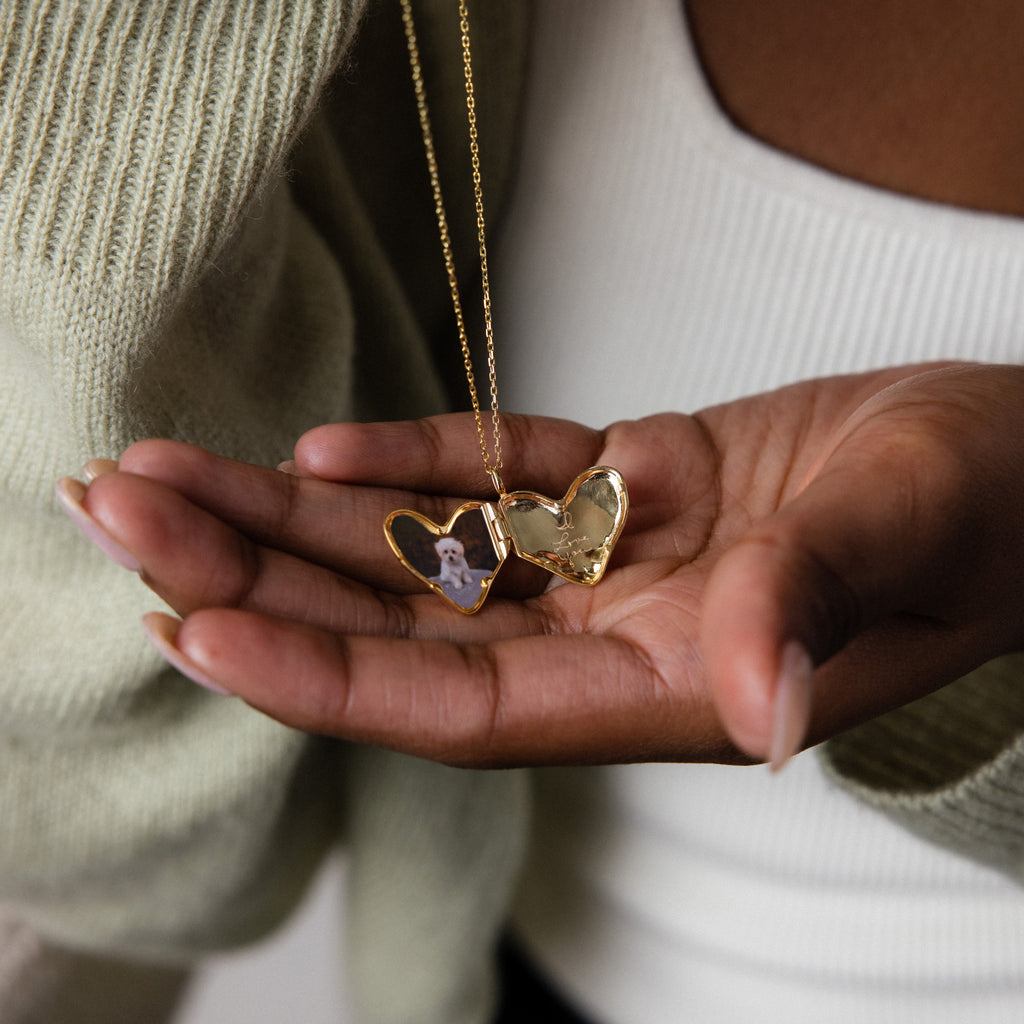 A hand holds an open Organic Heart Locket Necklace in 18K Gold with a small photo inside, displayed against a white top and green sweater—a timeless keepsake accessory.