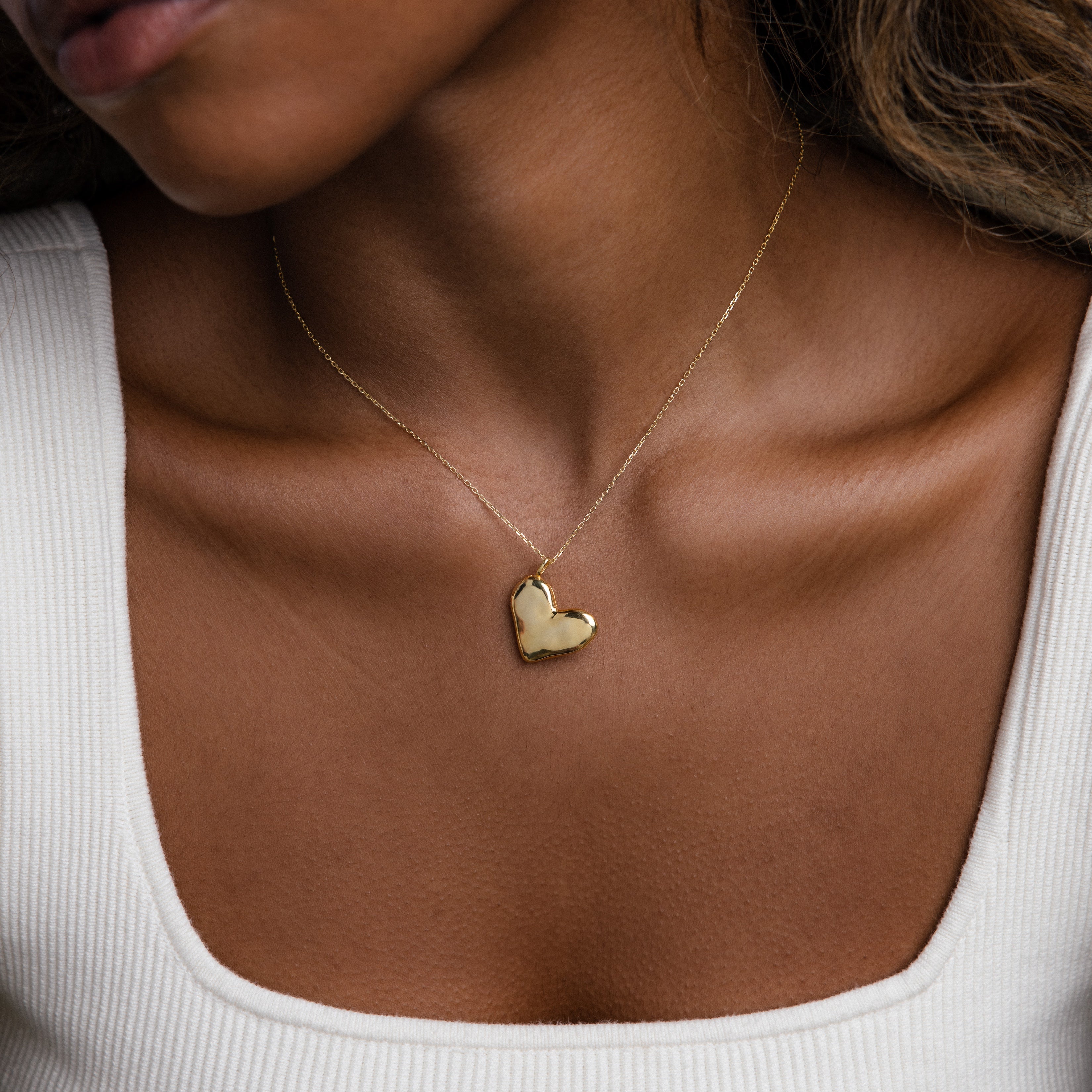 Woman wearing the Organic Heart Locket Necklace, a gold heart-shaped engraved locket, paired with a white square-neck top to showcase this timeless keepsake jewelry.