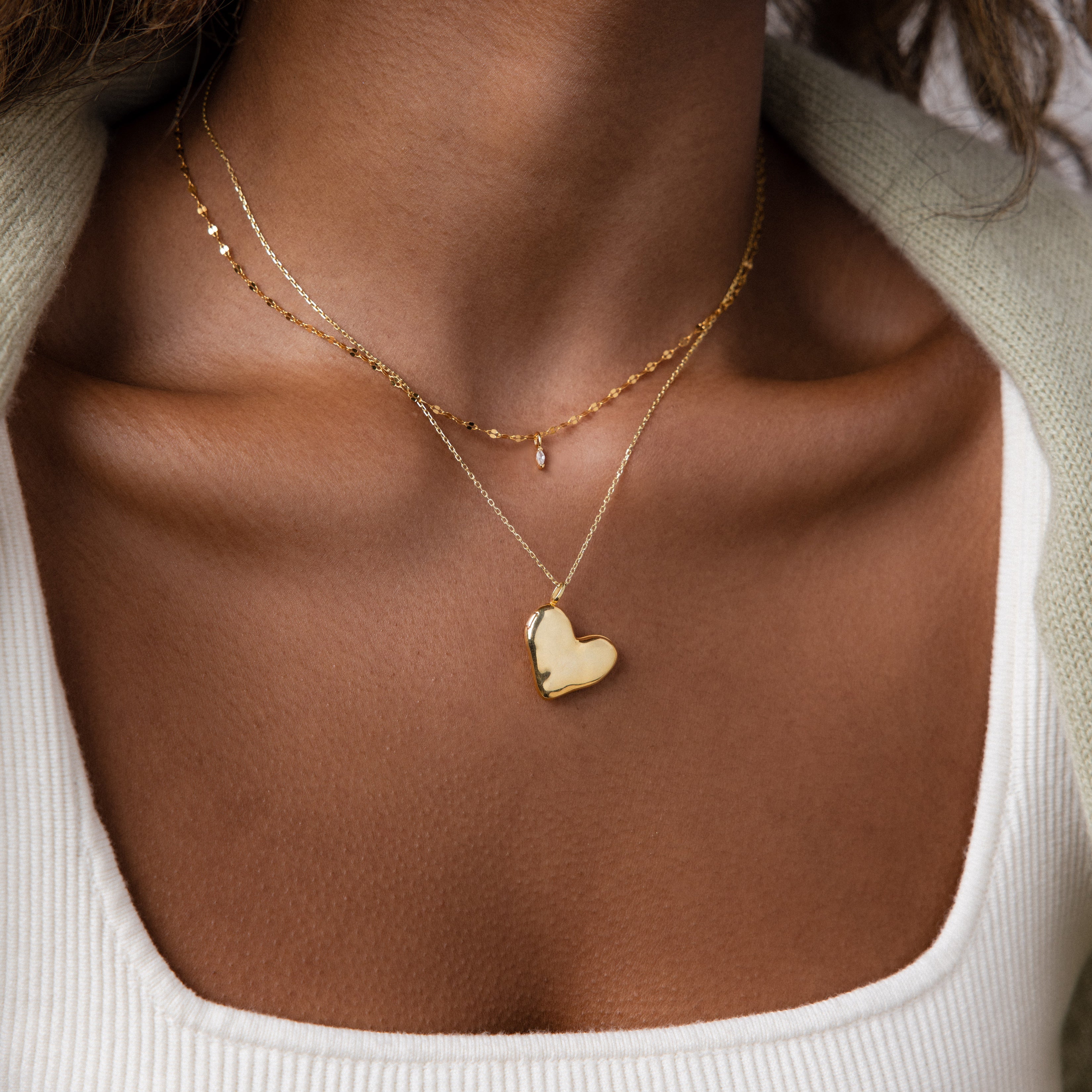 Close-up of a woman wearing two gold necklaces, one being the Organic Heart Locket Necklace in 18K Gold with a heart-shaped pendant, over a white top.