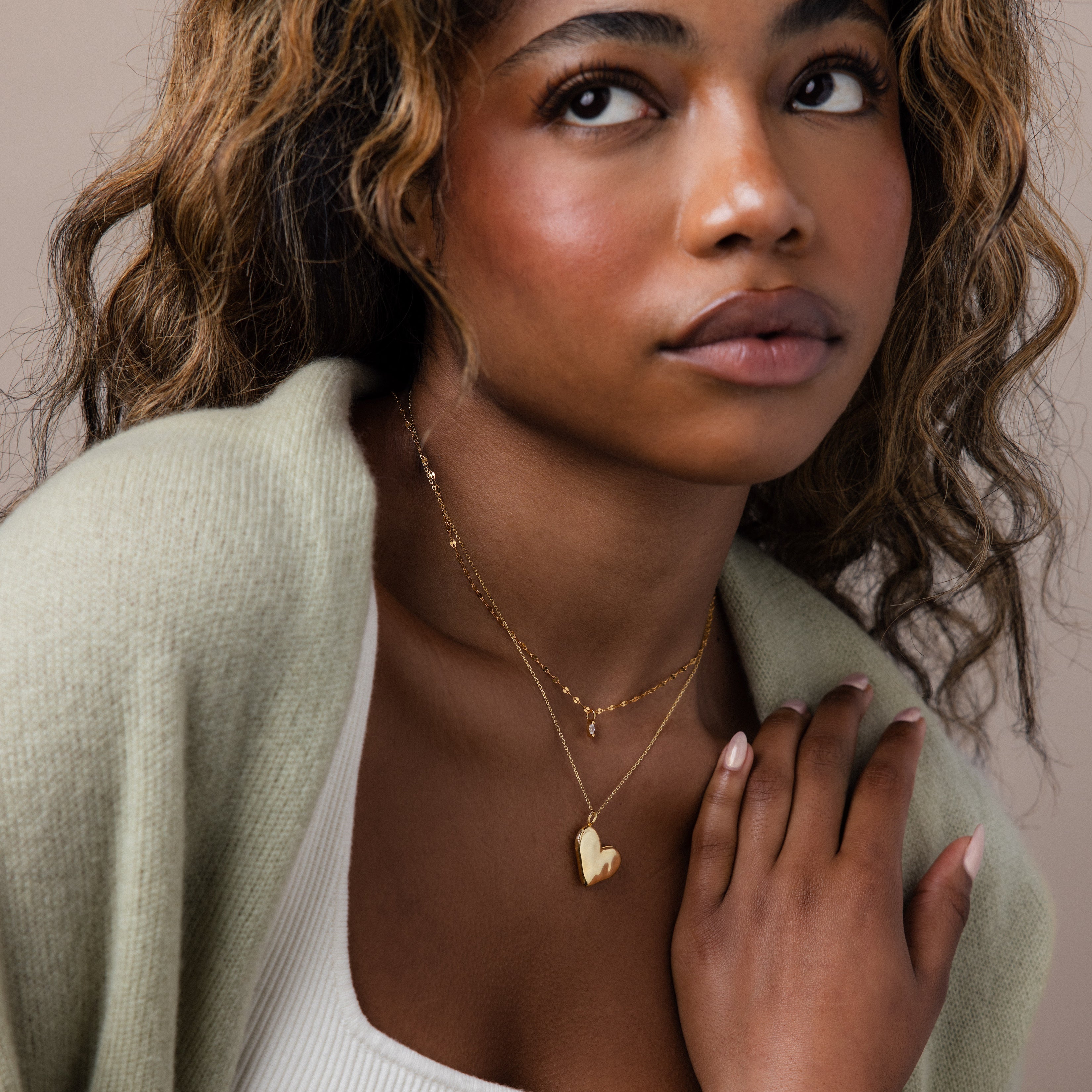A woman with wavy hair wears a light sweater and the Organic Heart Locket Necklace in 18K Gold, looking slightly upward. This beautiful keepsake adds elegance and personal charm to her style.