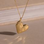 The Organic Heart Locket Necklace in 18K Gold features a heart-shaped locket on a delicate chain, shown against a softly blurred background.