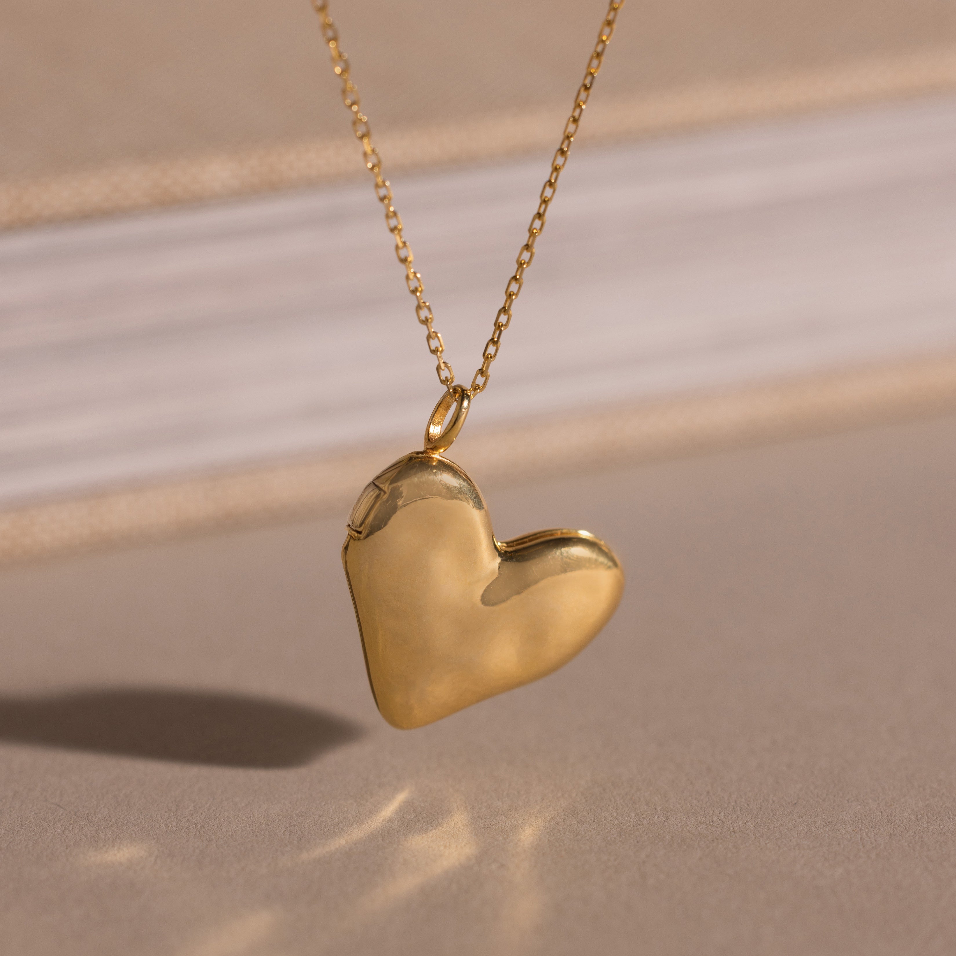 The Organic Heart Locket Necklace in 18K Gold features a heart-shaped locket on a delicate chain, shown against a softly blurred background.