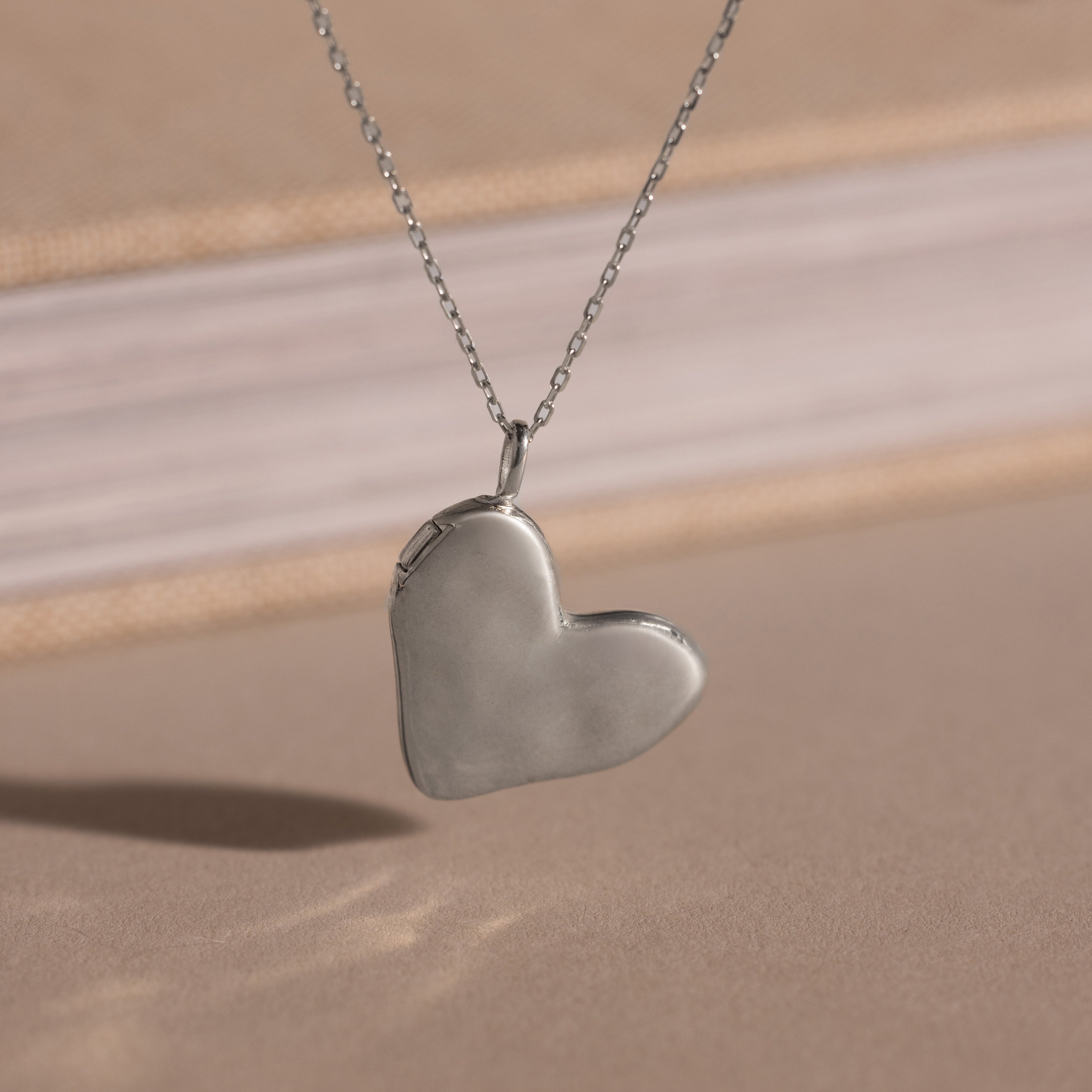 The Organic Heart Locket Necklace in Sterling Silver rests on a beige surface with a blurred book in the background, making it an ideal keepsake for preserving treasured memories.