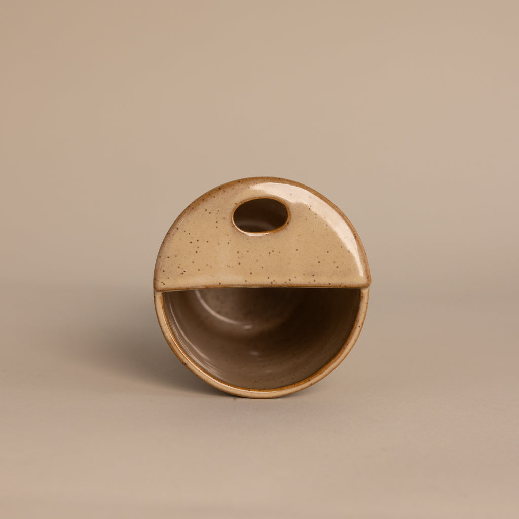 The Everyday Ceramic Mug is round and beige with a half-covered top, oval cutout handle, and subtle speckled glaze, shown on a neutral background.