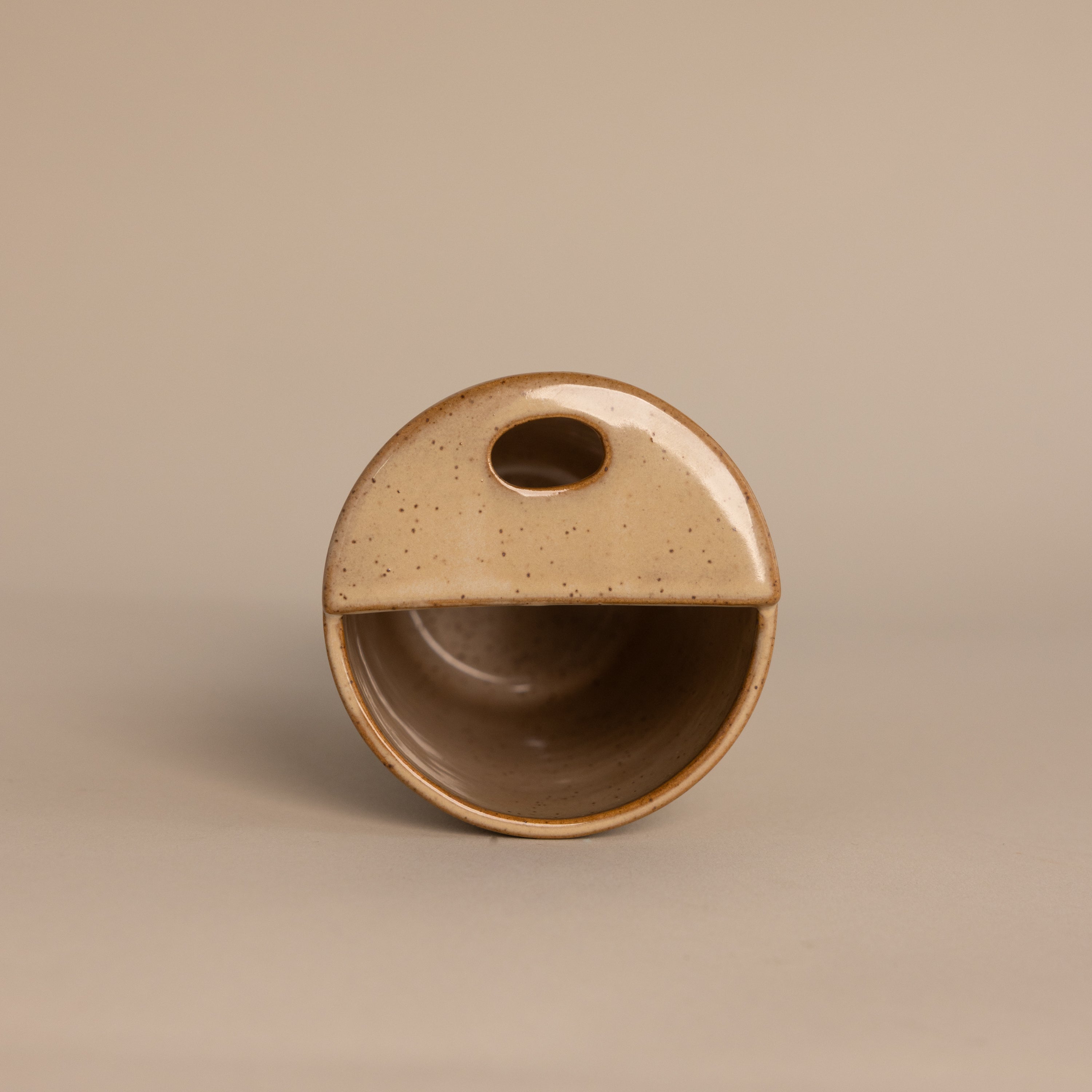 The Everyday Ceramic Mug is round and beige with a half-covered top, oval cutout handle, and subtle speckled glaze, shown on a neutral background.