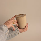 A hand in a denim sleeve holds the Everyday Ceramic Mug, featuring a beige speckled glaze and a small hole in the lid, set against a neutral background.