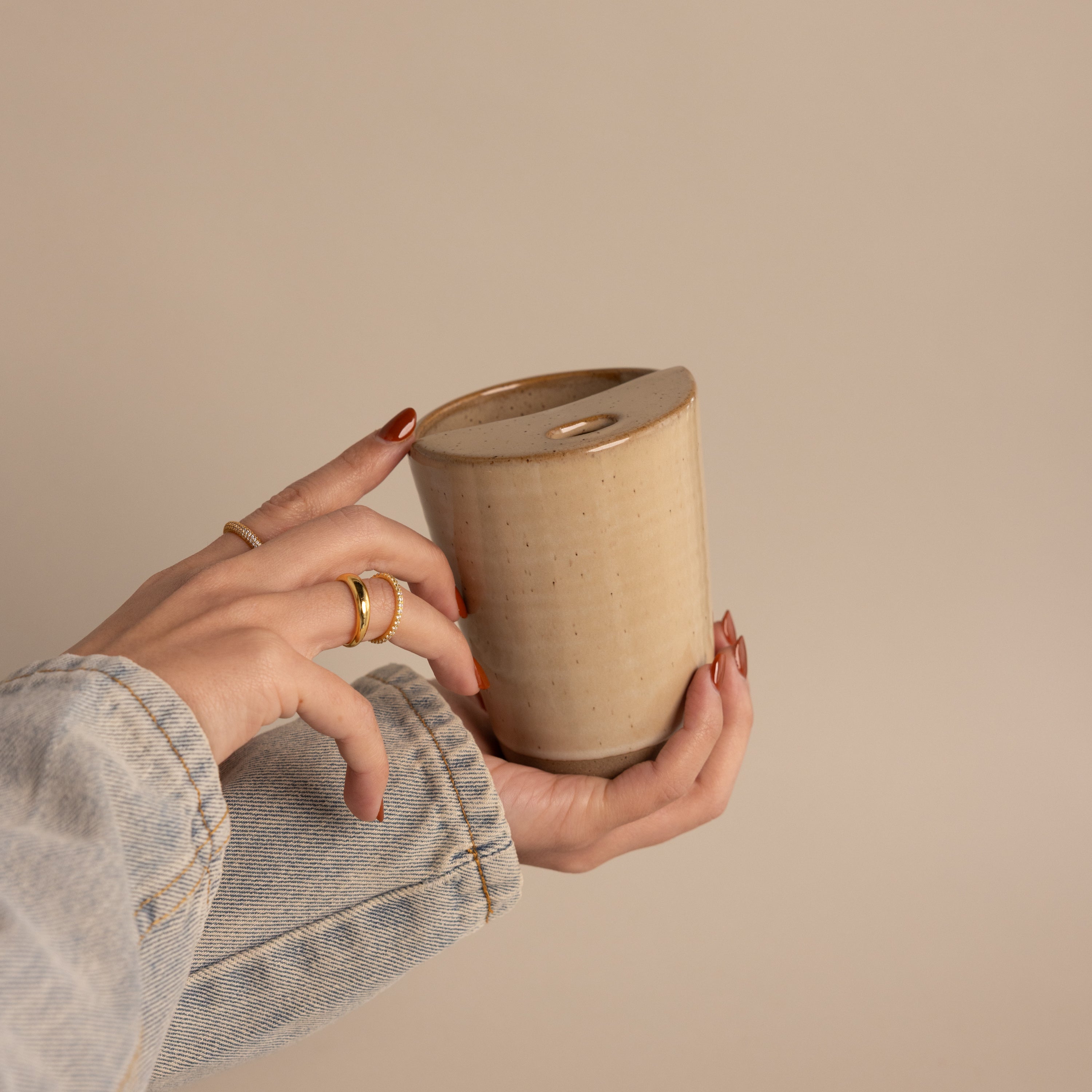 A hand in a denim sleeve holds the Everyday Ceramic Mug, featuring a beige speckled glaze and a small hole in the lid, set against a neutral background.