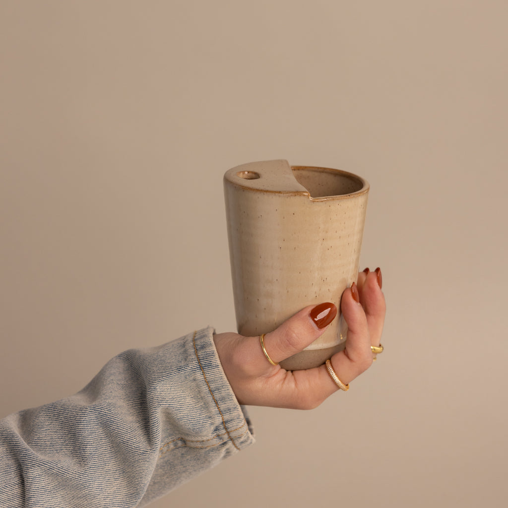A hand with painted nails holds the Everyday Ceramic Mug, which features a speckled glaze and a lid, set against a neutral background.