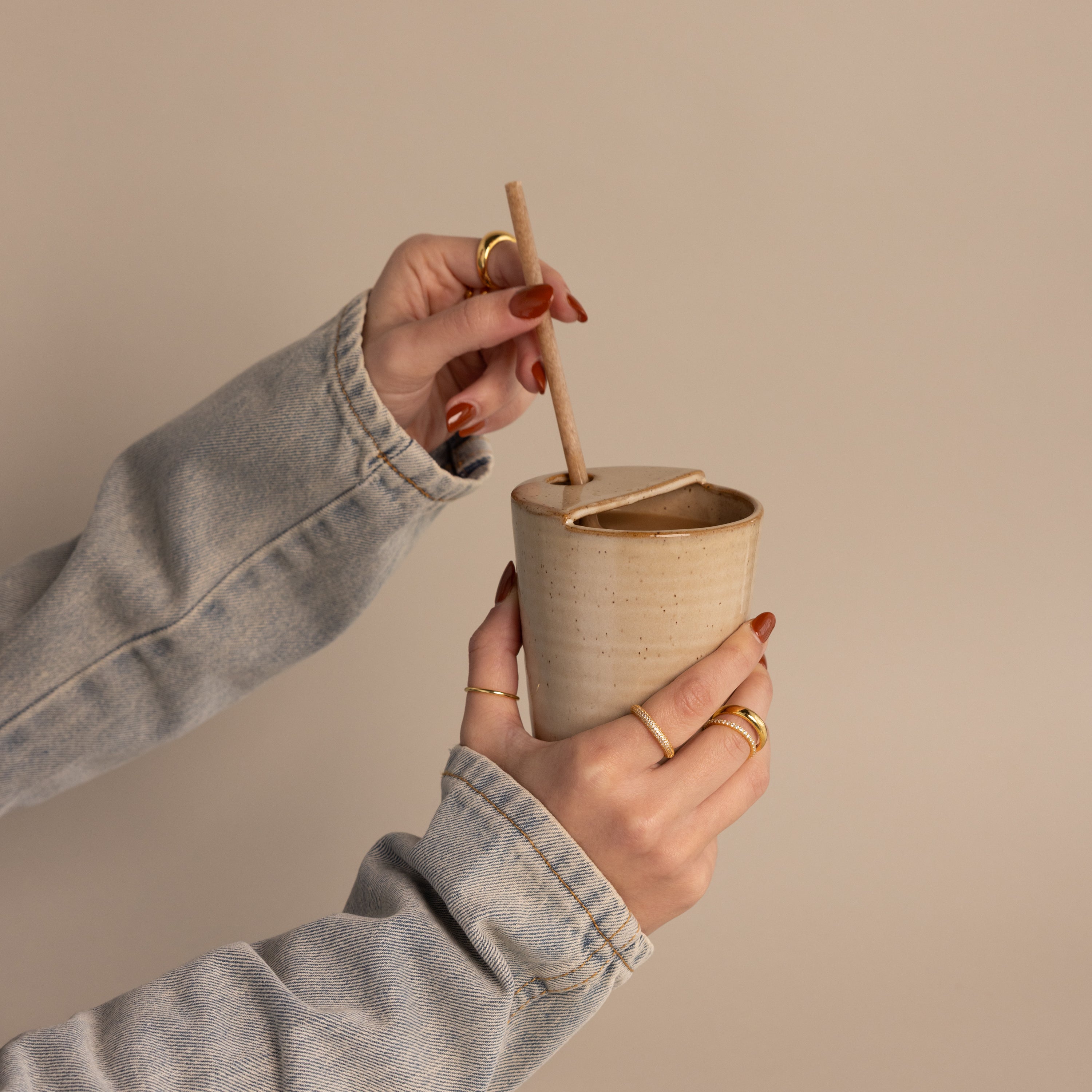 Hands stirring a drink in the Everyday Ceramic Mug, featuring a speckled glaze, while wearing rings and a denim jacket.