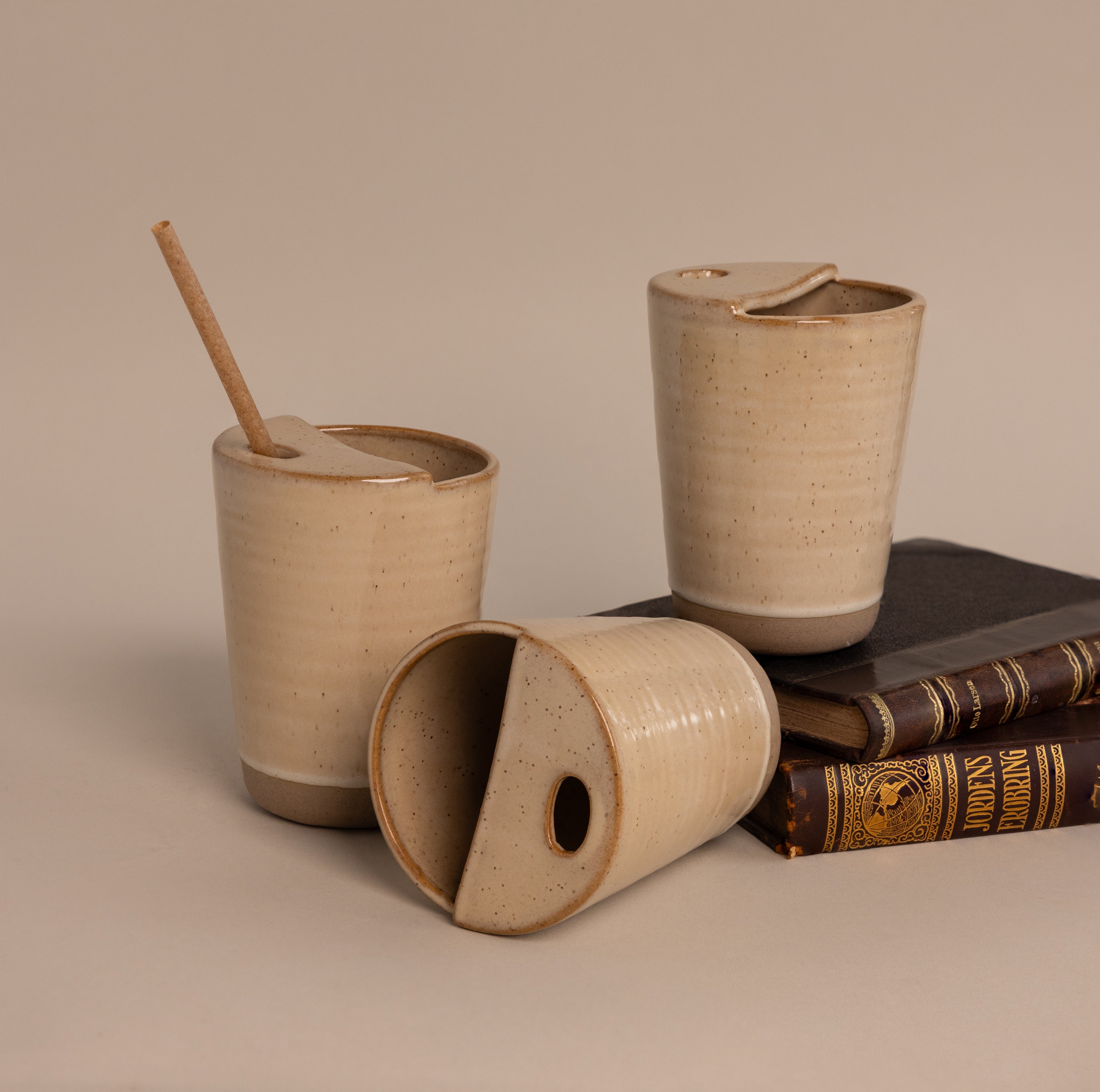 Three Everyday Ceramic Mugs with lids—including one with a straw and a speckled glaze—are displayed on and next to vintage books against a beige background.