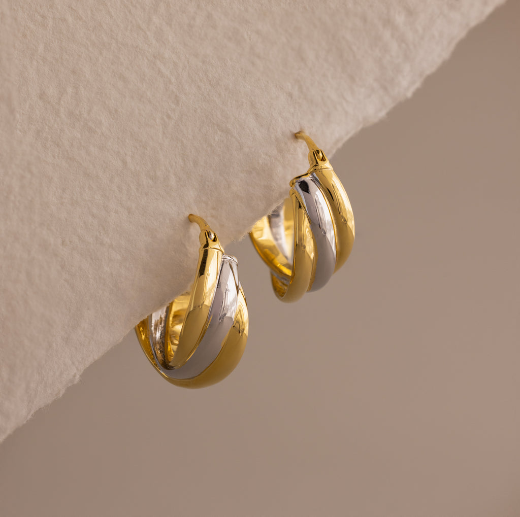 The Intertwined Mixed Metal Hoops are gold and silver twisted hoop earrings, displayed on a textured off-white surface.