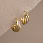 The Intertwined Mixed Metal Hoops are gold and silver twisted hoop earrings, displayed on a textured off-white surface.