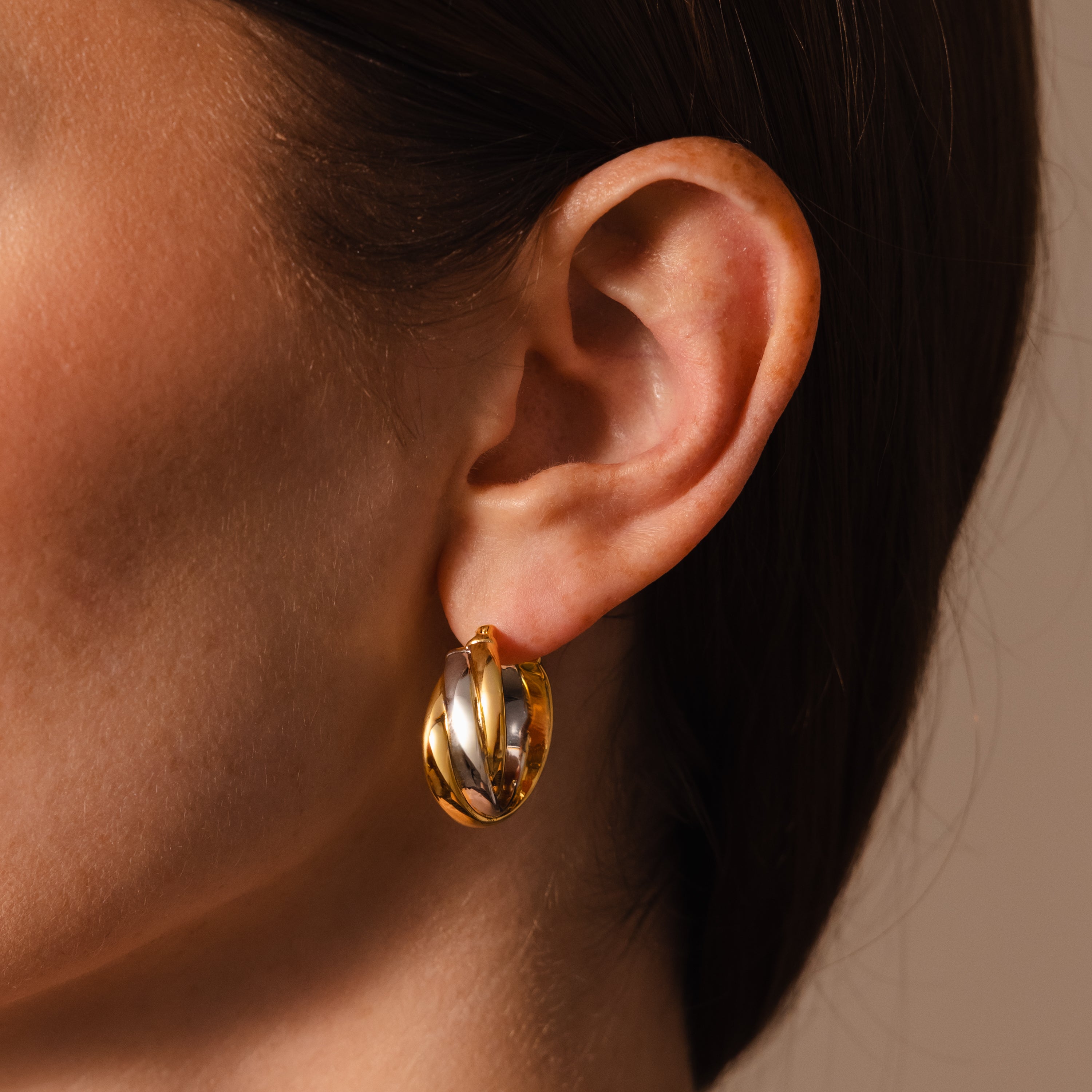 Close-up of a person’s ear adorned with Intertwined Mixed Metal Hoops in gold and silver, highlighting their elegant shine against a neutral background.