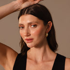A woman with long brown hair, wearing Intertwined Mixed Metal Hoops and a black top, poses with one arm raised against a neutral background.