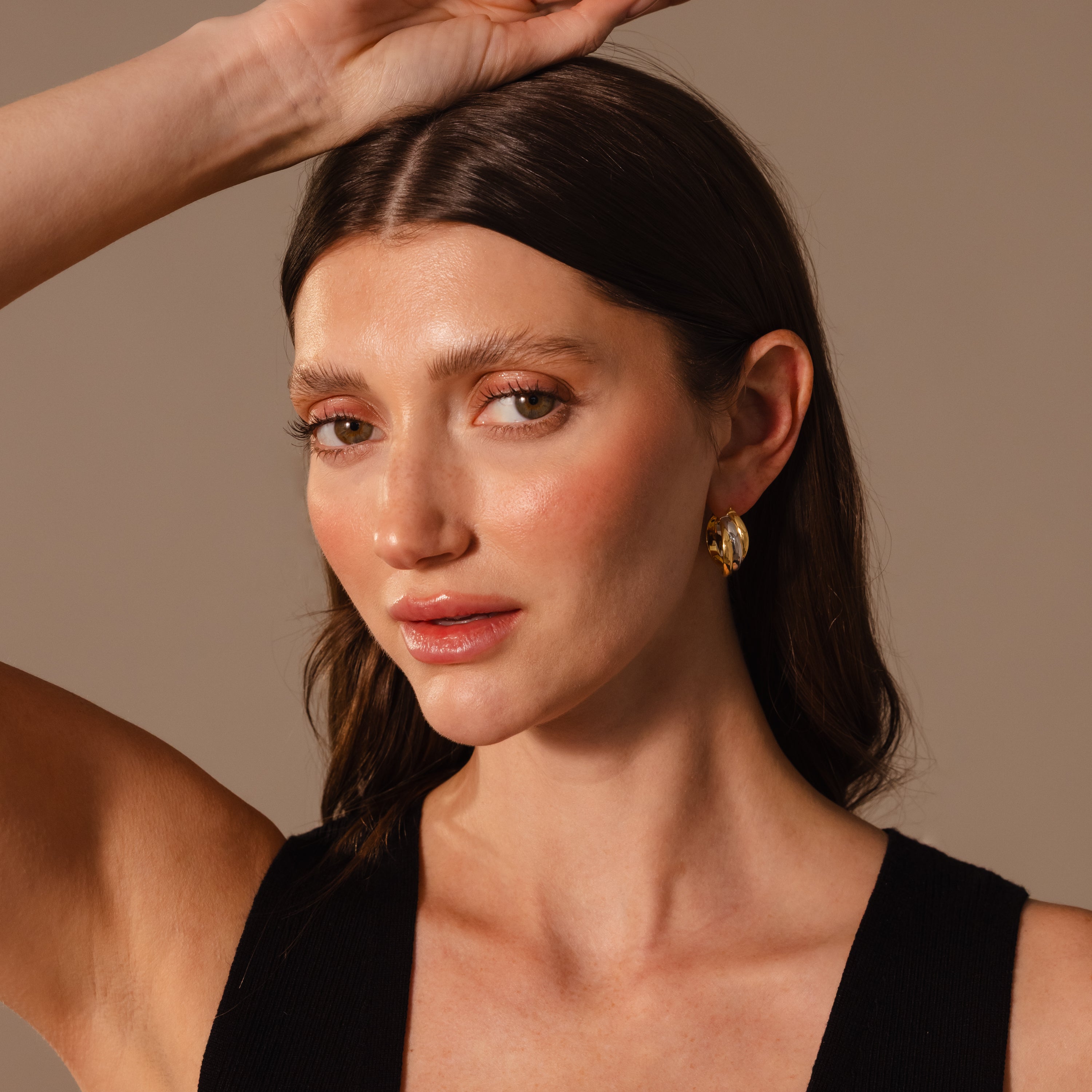 A woman with long brown hair, wearing Intertwined Mixed Metal Hoops and a black top, poses with one arm raised against a neutral background.