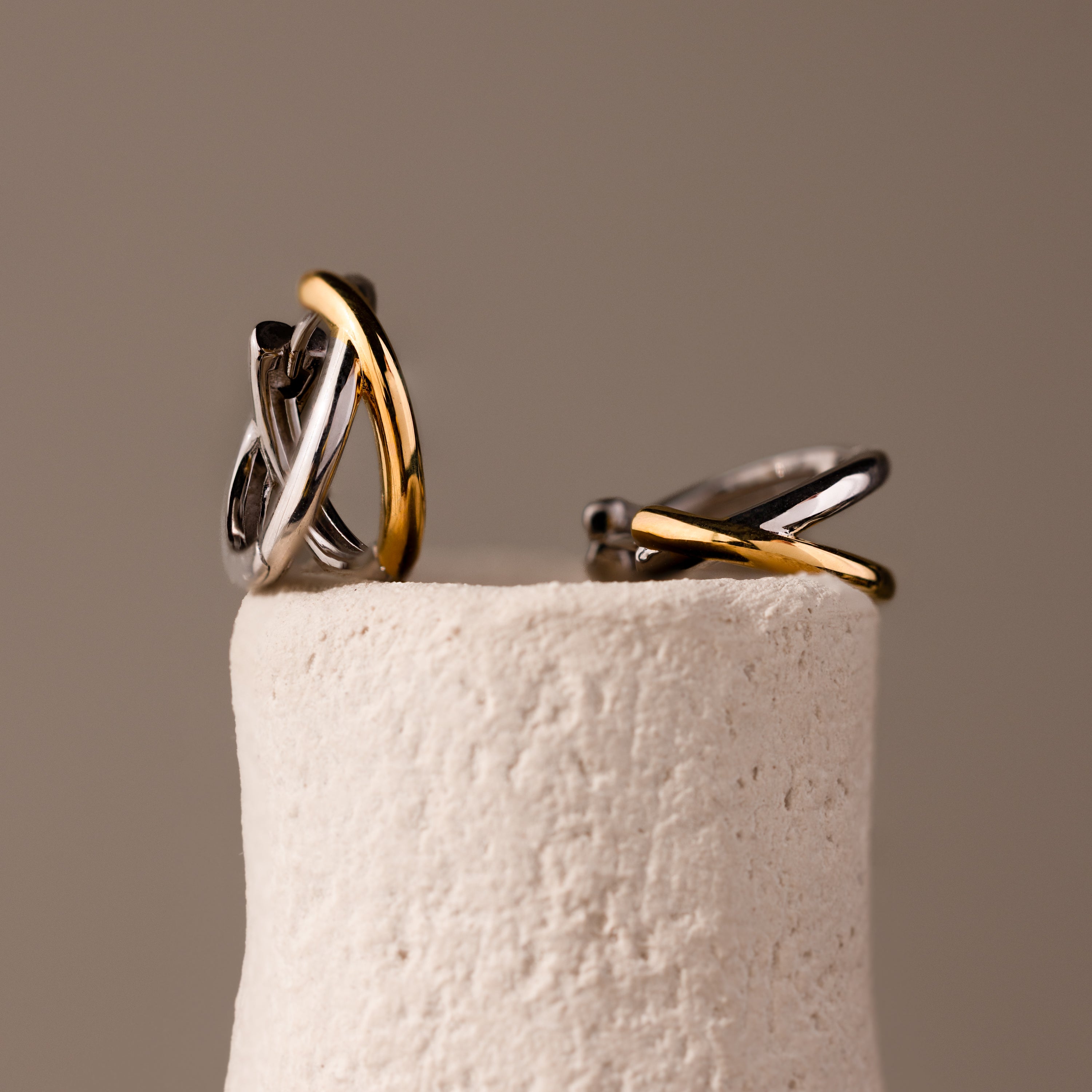 Laced Mixed Metal Huggies, featuring intertwined silver and gold in a sculptural crossover design, are showcased on a textured off-white cylindrical stand.
