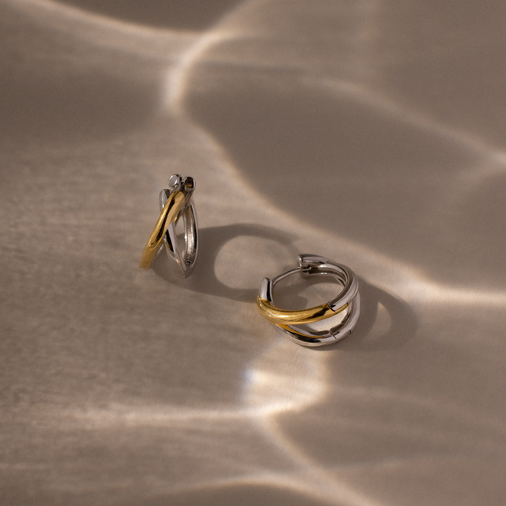 The Laced Mixed Metal Huggies, featuring gold and silver tones, are displayed on a beige surface with wavy light reflections, highlighting their sculptural crossover design.