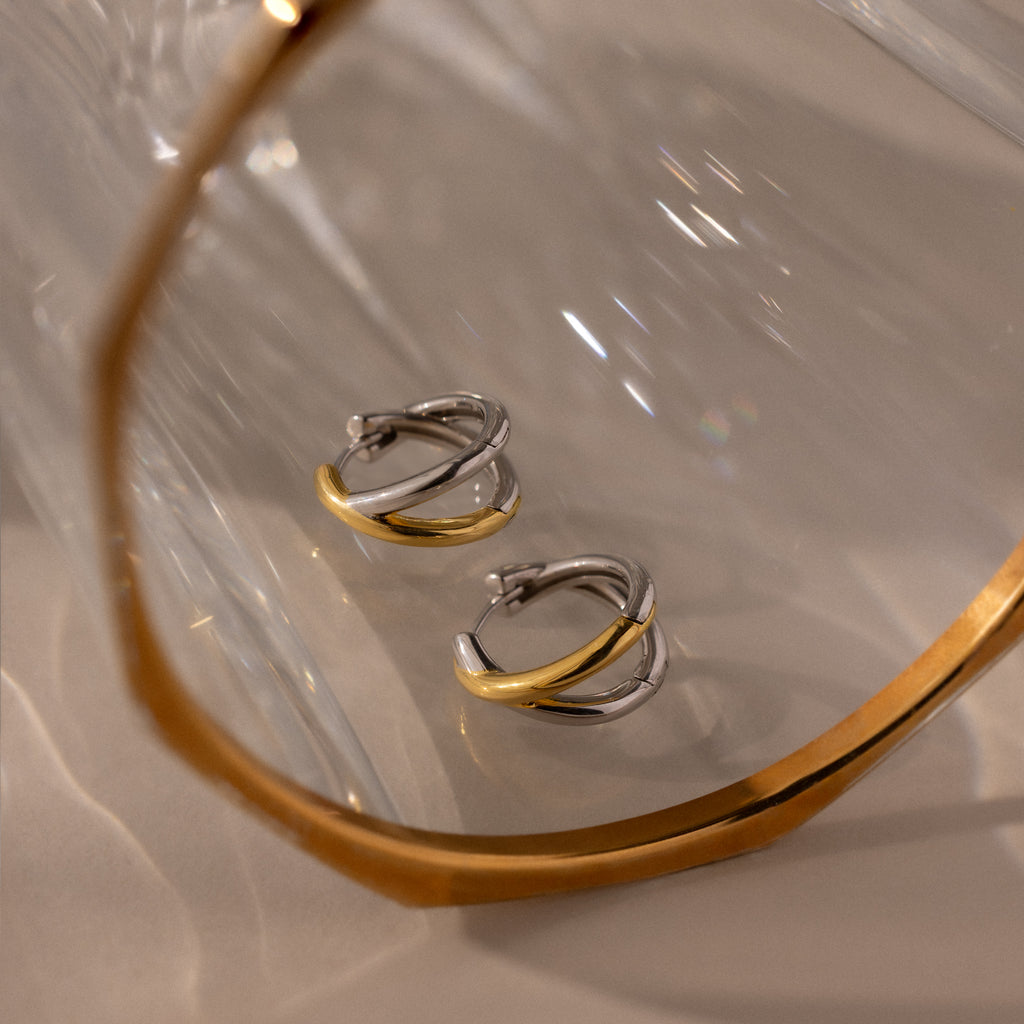 Laced Mixed Metal Huggies—two-tone gold and silver earrings—are reflected in a glass surface with a gold-edged frame, highlighting their sculptural crossover design.