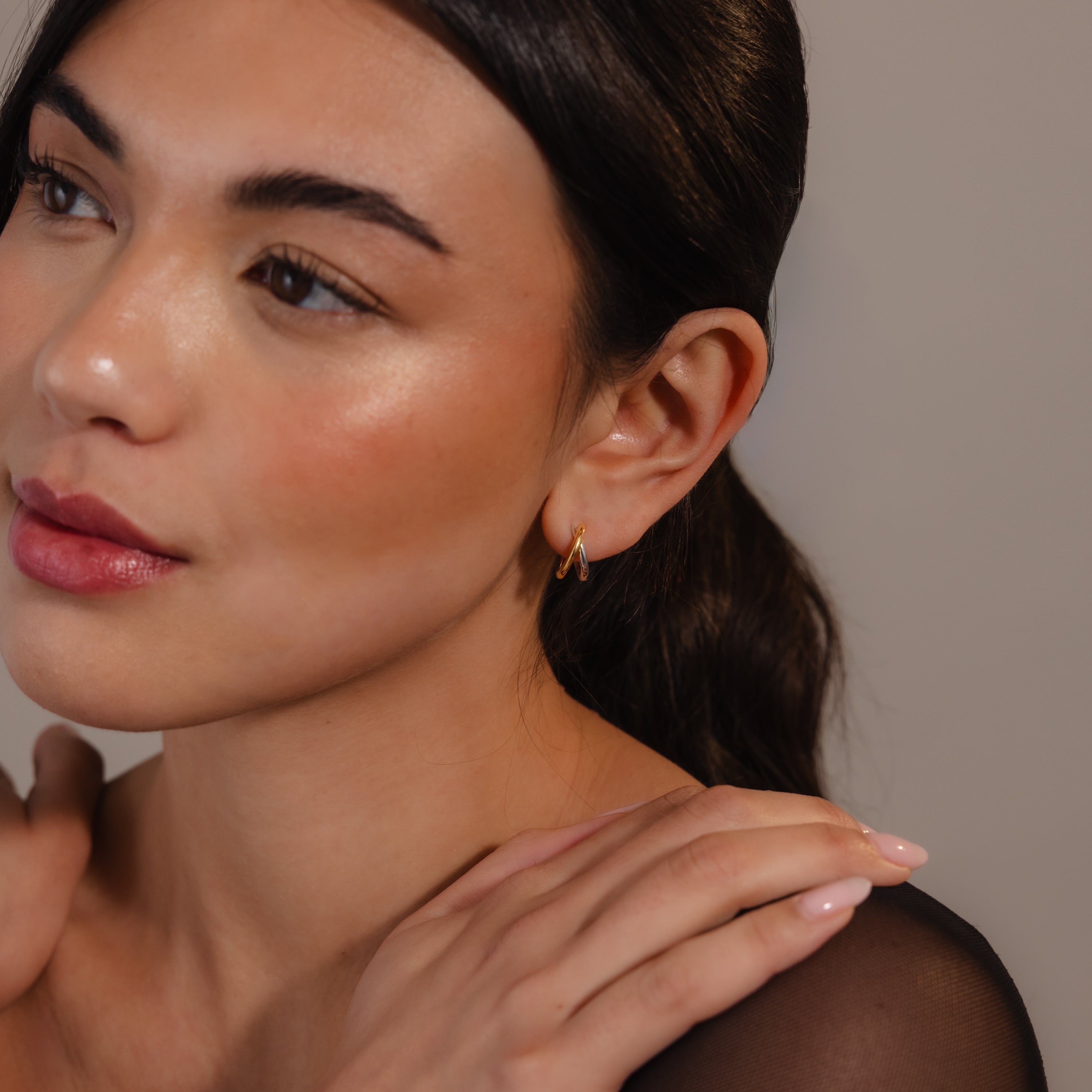 Woman with dark hair in a ponytail, wearing Laced Mixed Metal Huggies and a sheer black top, looking to the side.