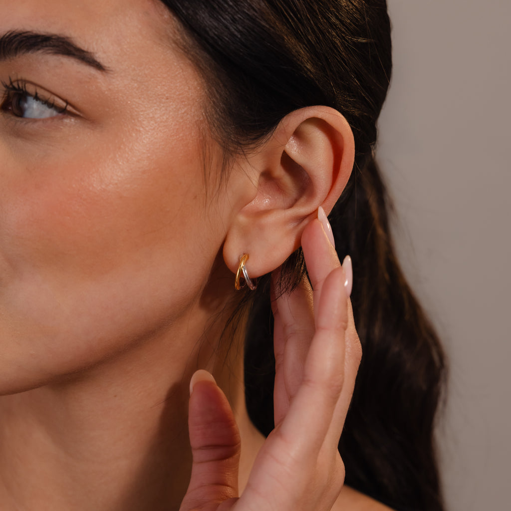 A woman with long dark hair touches her ear, wearing Laced Mixed Metal Huggies in gold and silver.
