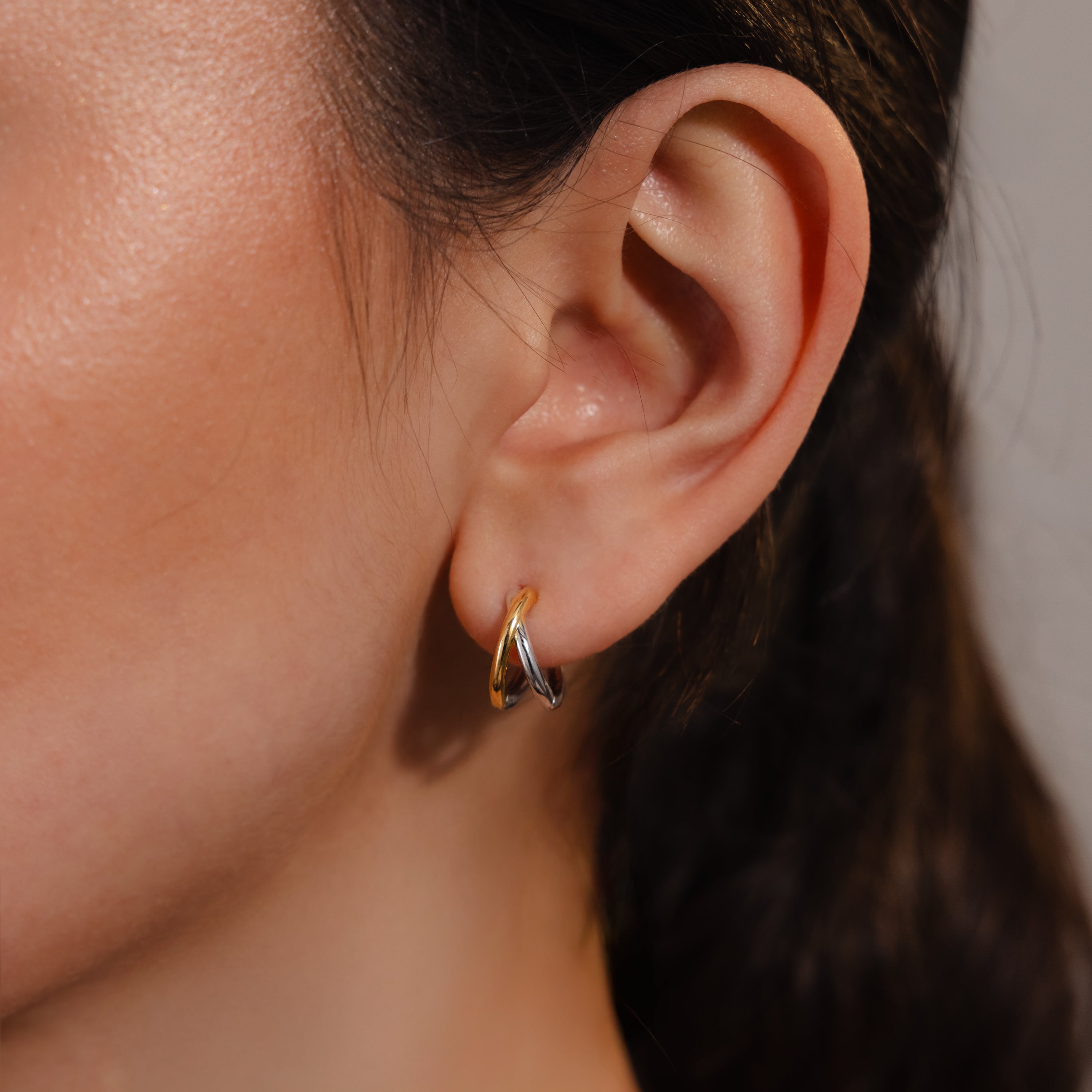 Close-up of a woman’s ear wearing Laced Mixed Metal Huggies—gold and silver earrings with a sculptural, twisted hoop crossover design.