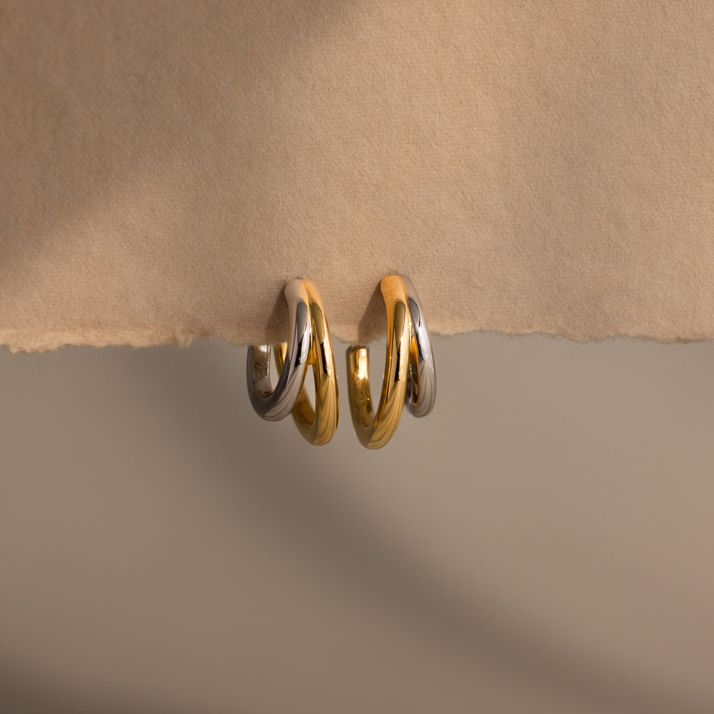 Two pairs of Mixed Metal Duo Hoops—featuring gold and silver—rest elegantly on beige paper, highlighting their modern hoop earring design.