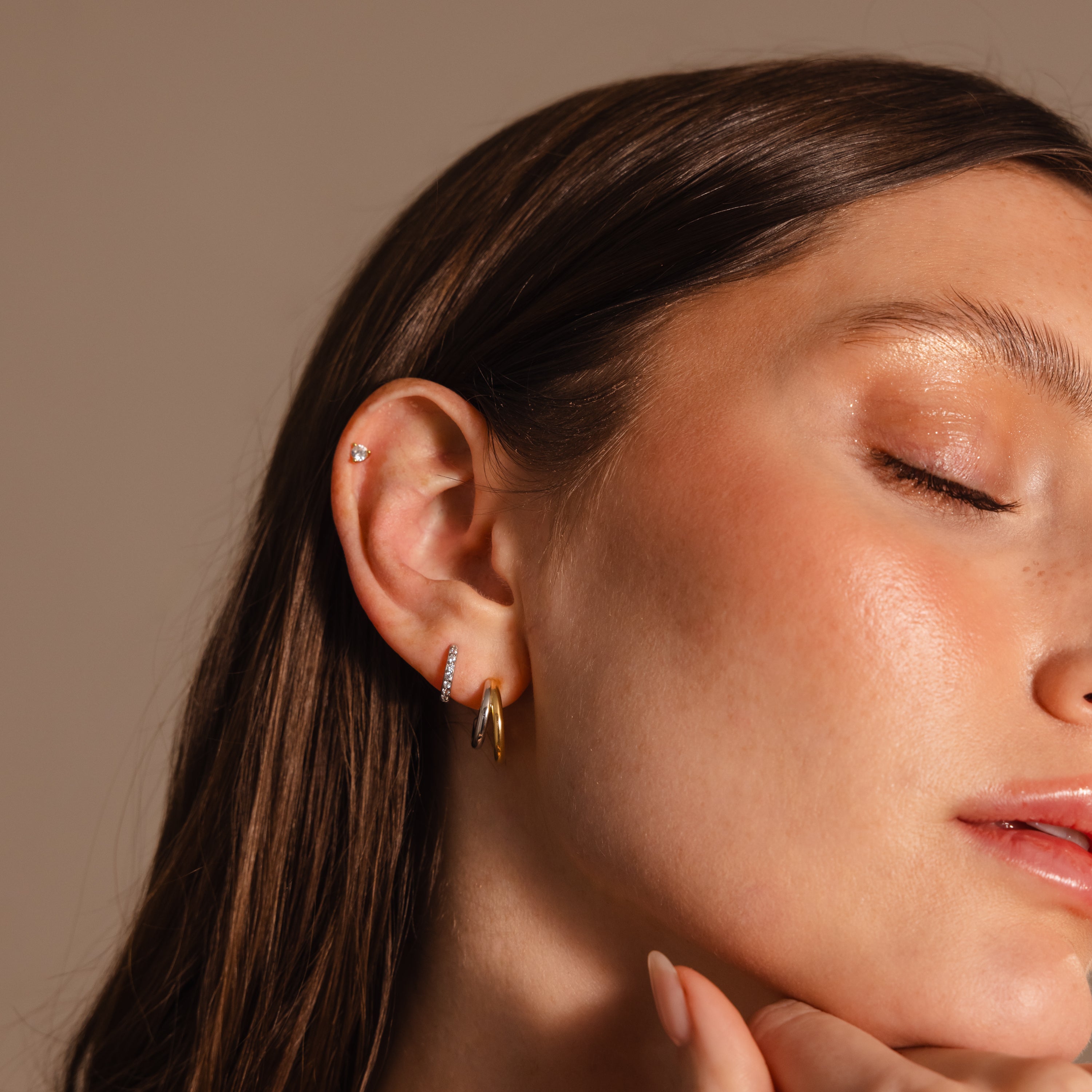 A woman with closed eyes gently touches her chin, showcasing the Mixed Metal Duo Hoops against a neutral background.