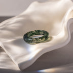 The Skinny Agate Ring, featuring translucent green marbling resin, is displayed on a wavy white ceramic dish in soft natural light—an eye-catching accessory ideal for gemstone lovers.