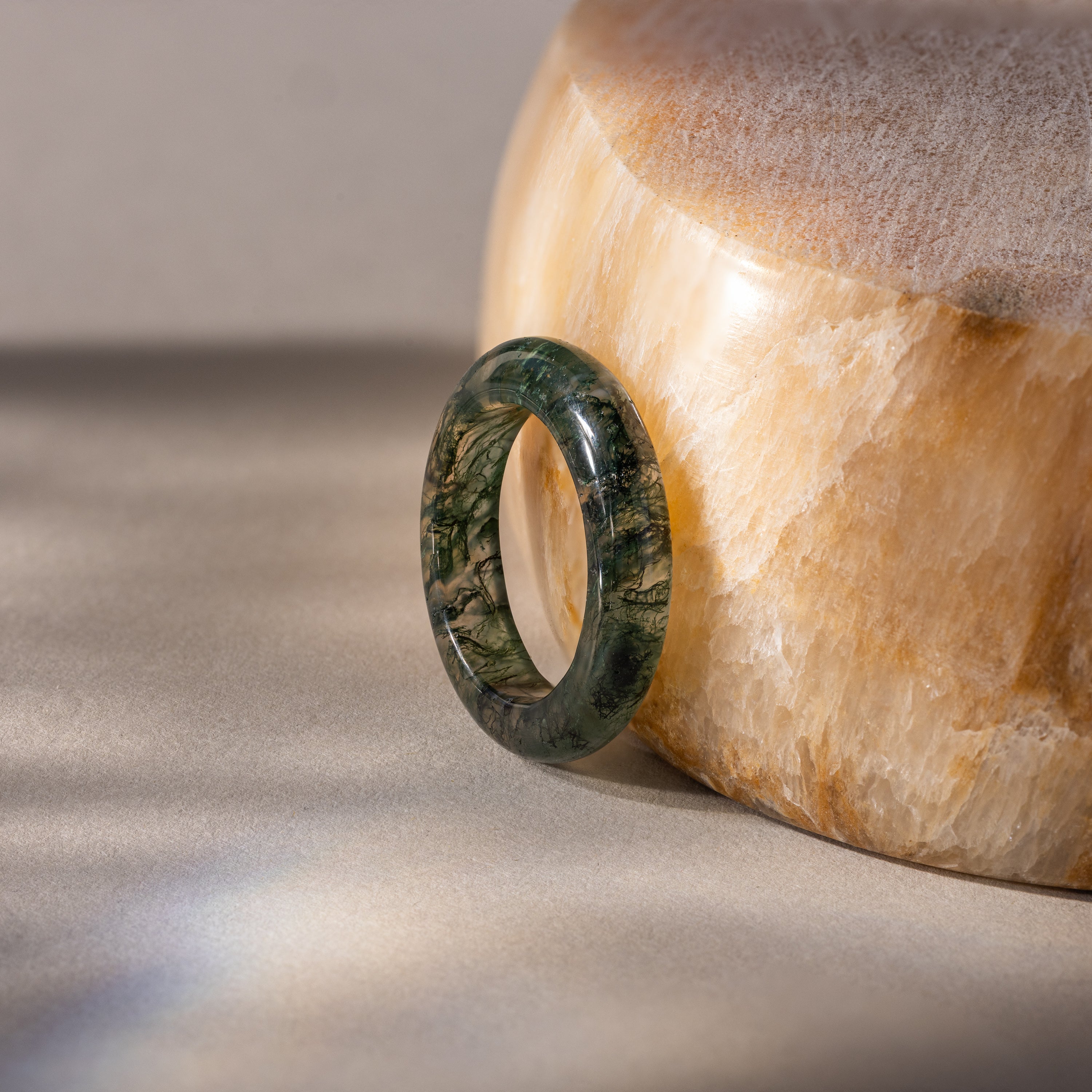 The Skinny Agate Ring, featuring green marbling, stands upright against a polished light brown stone—an ideal choice for gemstone lovers drawn to natural elegance.