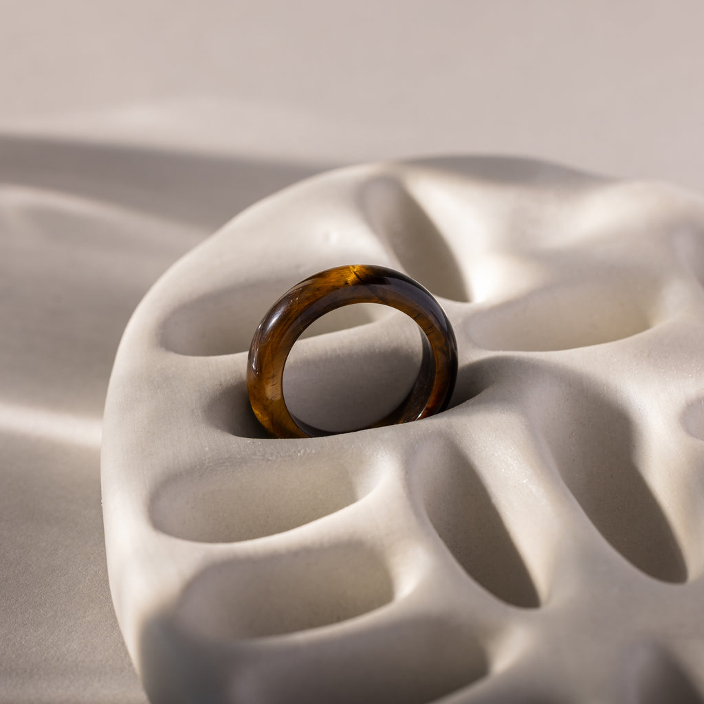 A Skinny Tiger's Eye Ring in brown translucent hues rests on a sculpted white ceramic surface with organic curves, highlighting the earthy beauty of this unique jewelry piece.