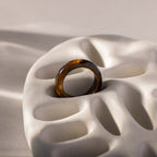 A Skinny Tiger's Eye Ring in brown translucent hues rests on a sculpted white ceramic surface with organic curves, highlighting the earthy beauty of this unique jewelry piece.