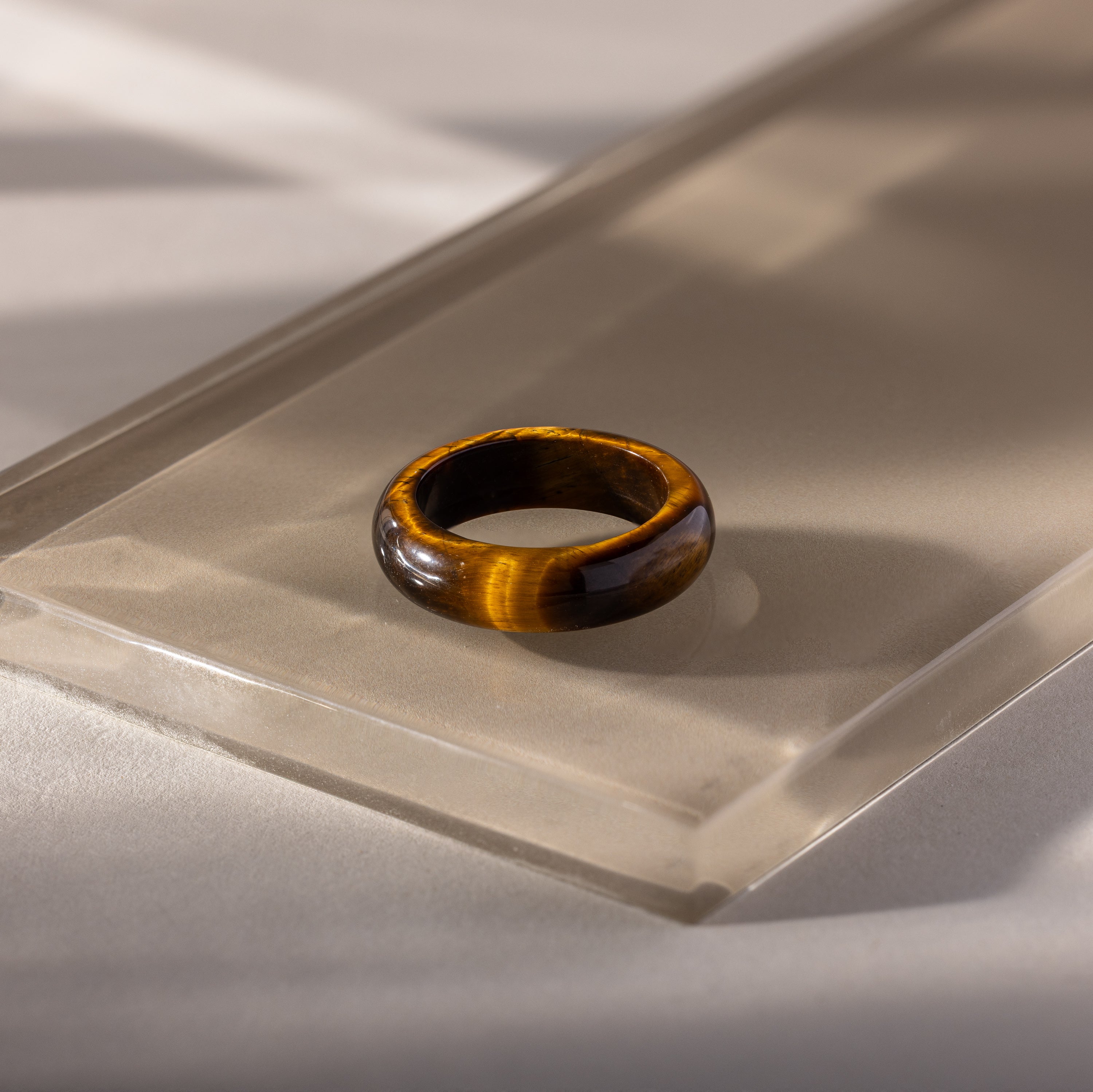 The Skinny Tiger's Eye Ring, ideal for stacking, is displayed on a clear acrylic block and illuminated by natural light—an elegant, earthy accessory.