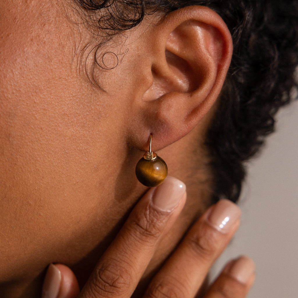 Close-up of a person wearing Tiger's Eye Dome Drop Earrings in 18K Gold, with a hand touching their neck near the earlobe, highlighting the elegant natural stone dome drop design.