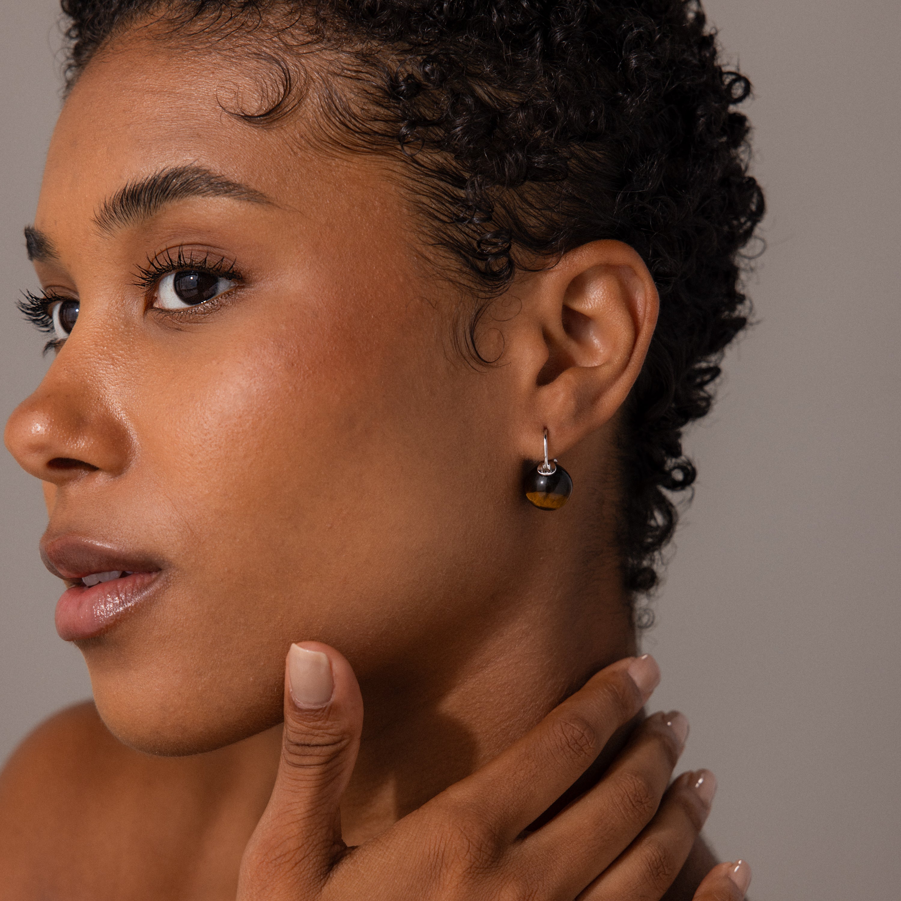 A woman with short curly hair wears Tiger's Eye Dome Drop Earrings in Sterling Silver, touches her neck, and looks to the side.