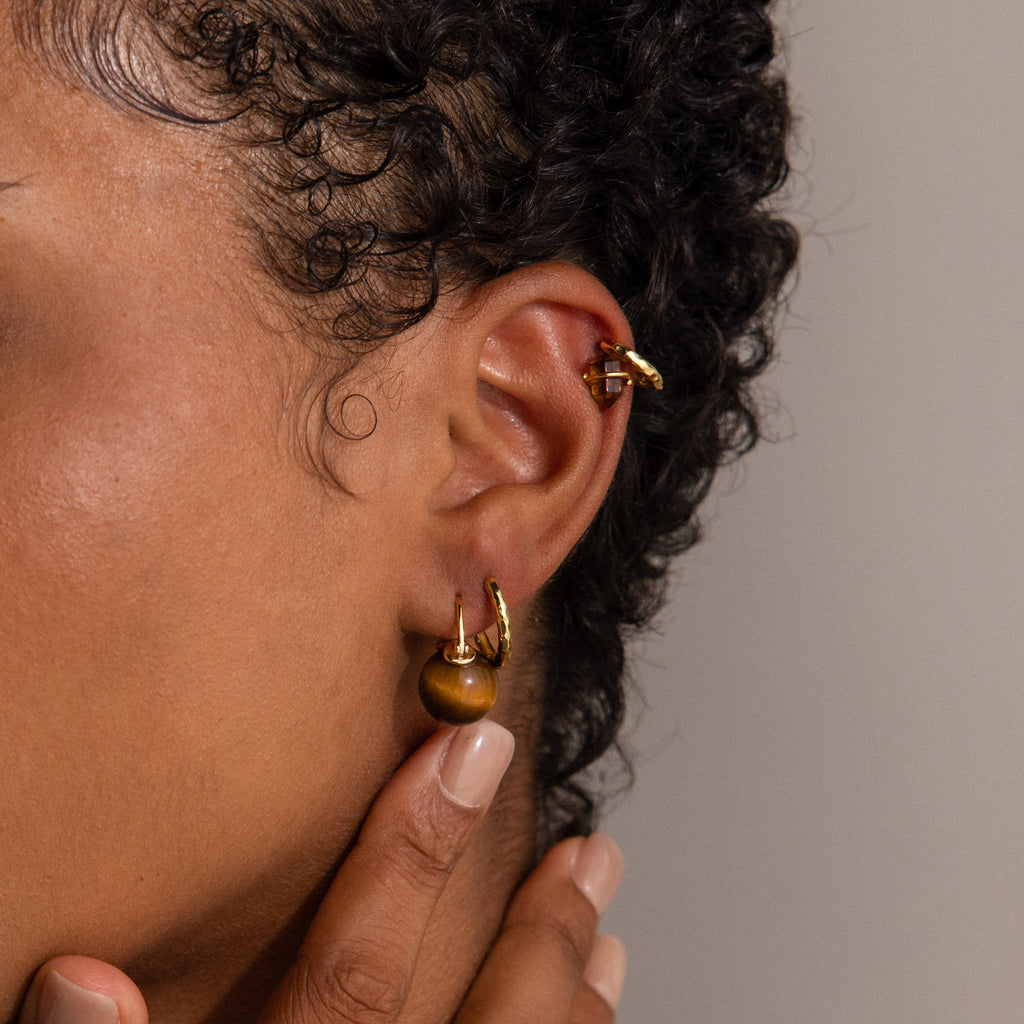 A person with curly hair wears Tiger's Eye Dome Drop Earrings in 18K Gold, touching their ear with one hand.