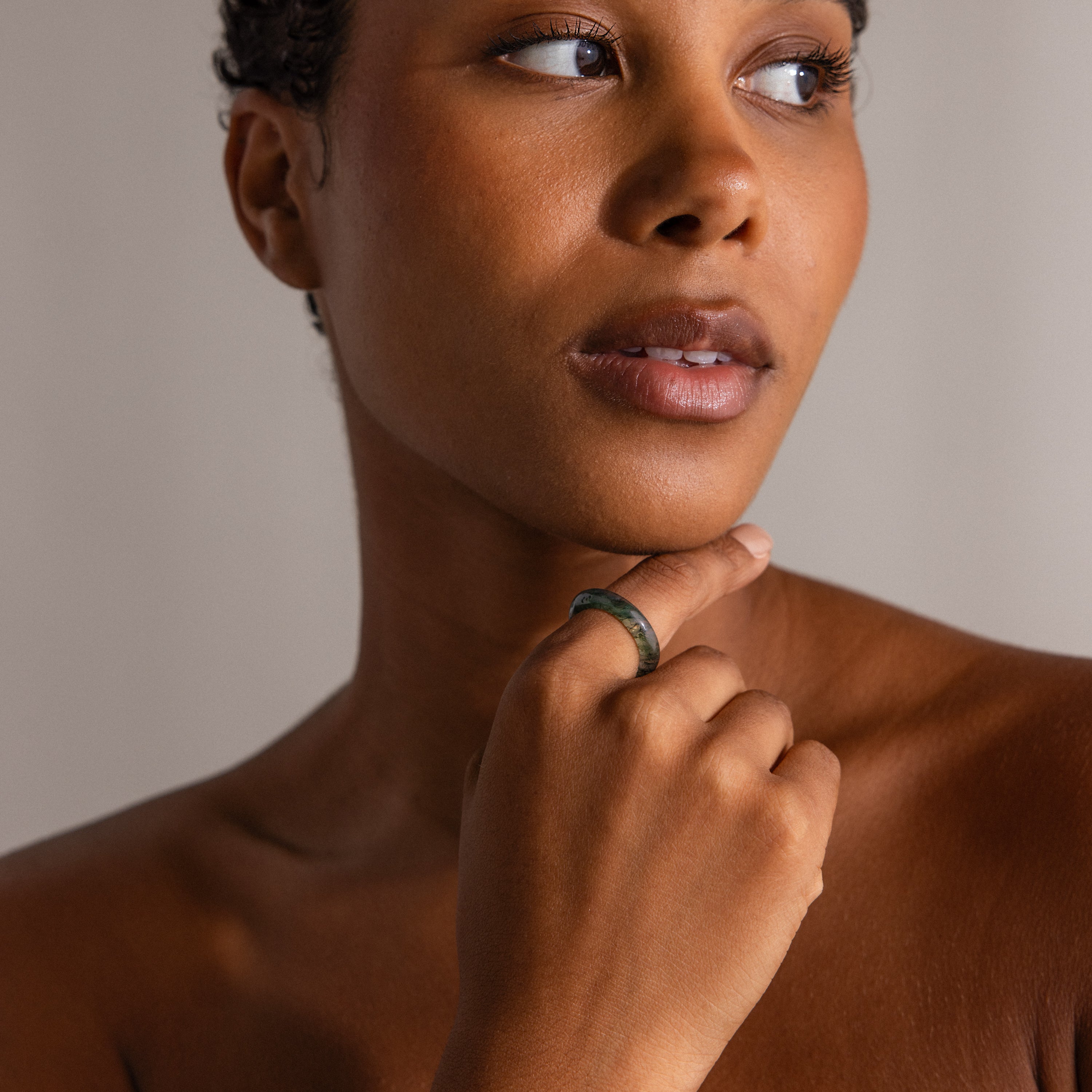 A woman with short hair gazes to the side, touching her chin while wearing the Skinny Agate Ring—an ideal choice for gemstone lovers seeking unique green marbling.