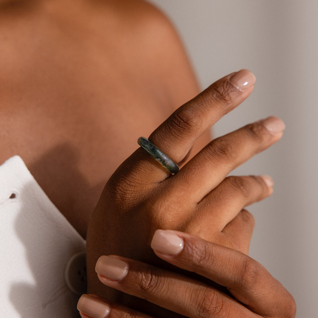 A hand with nude polish wears the Skinny Agate Ring, showcasing its striking green marbling as it rests on the opposite shoulder—a stunning choice for gemstone enthusiasts.