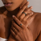 A woman with natural nails wears multiple rings, including the Skinny Agate Ring, featuring green marbling, as she touches her neck and collarbone—ideal for gemstone enthusiasts.