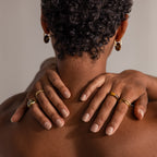 A person with short curly hair shows their back, hands on neck, wearing the Skinny Tiger's Eye Ring and gold hoop earrings—an effortless showcase of earthy jewelry.
