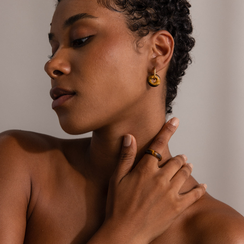 A woman with short curly hair wears gold hoop earrings and the Skinny Tiger's Eye Ring, touching her neck against a neutral background.