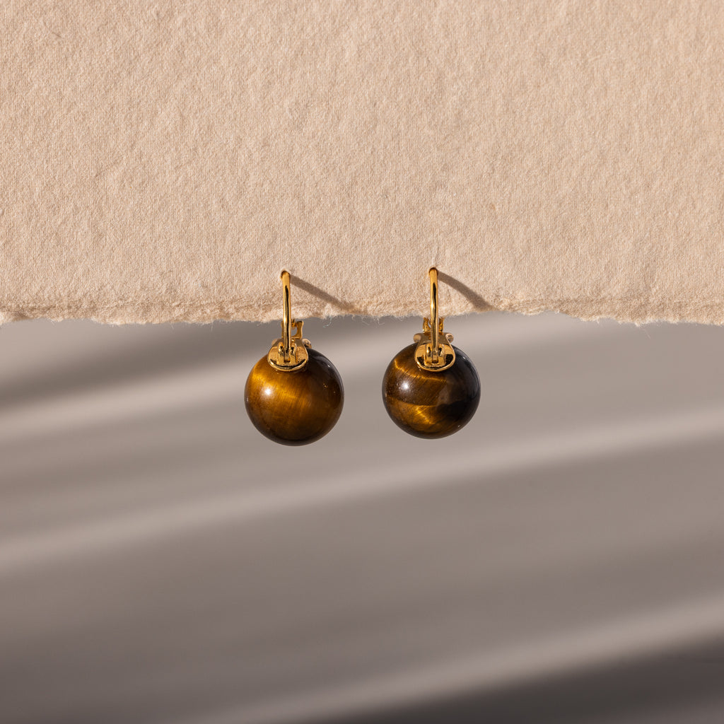 Tiger's Eye Dome Drop Earrings in 18K Gold, featuring round brown stones, are displayed gracefully on a textured beige surface and bathed in soft, natural light.