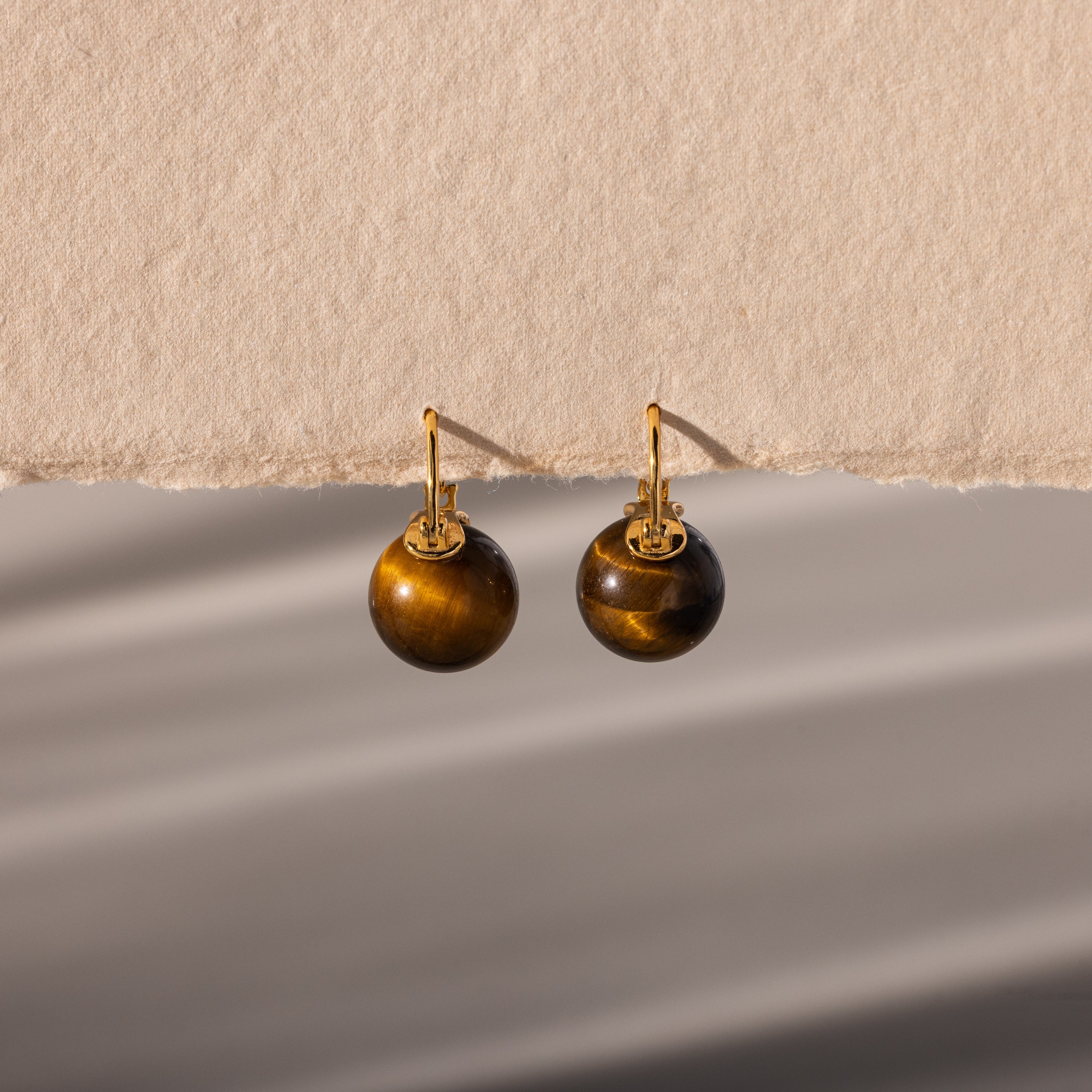 Tiger's Eye Dome Drop Earrings in 18K Gold, featuring round brown stones, are displayed gracefully on a textured beige surface and bathed in soft, natural light.
