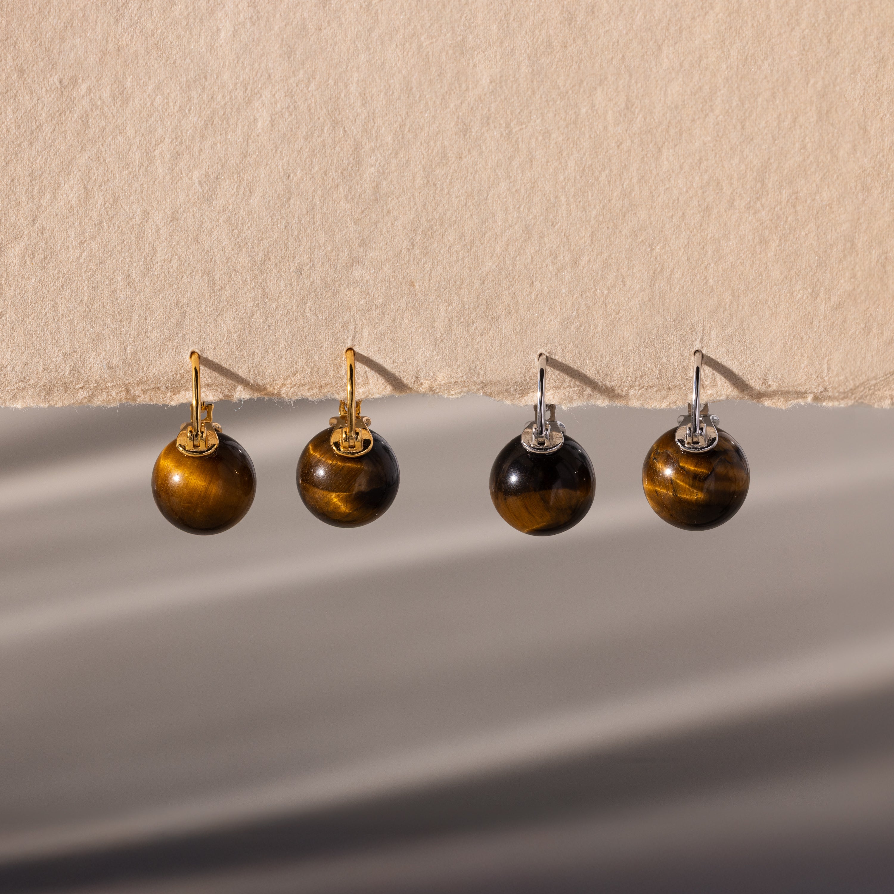 Two pairs of Tiger's Eye Dome Drop Earrings—crafted with natural stone and featuring gold or silver hooks—are elegantly displayed on a beige surface.