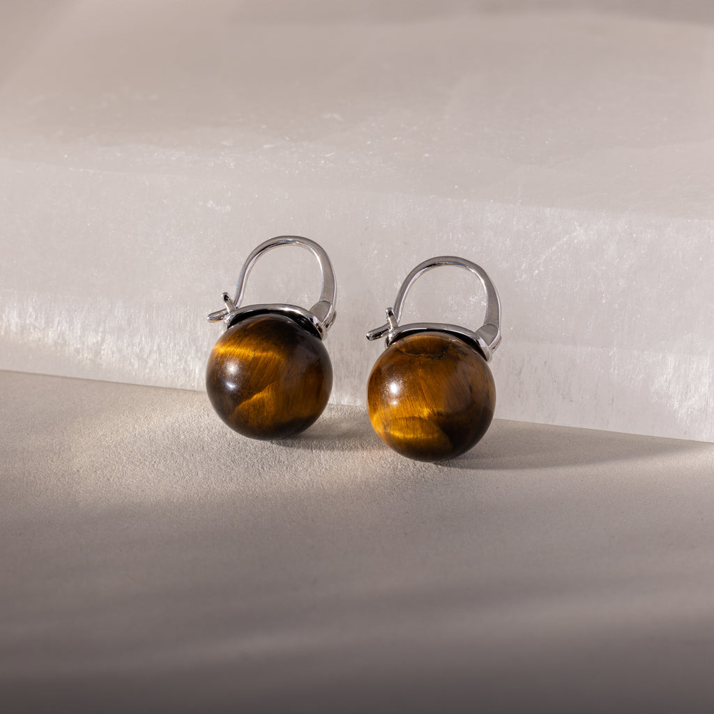 Tiger's Eye Dome Drop Earrings in Sterling Silver, featuring natural stone, are elegantly displayed on a light stone surface with soft lighting.