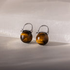 Tiger's Eye Dome Drop Earrings in Sterling Silver, featuring natural stone, are elegantly displayed on a light stone surface with soft lighting.