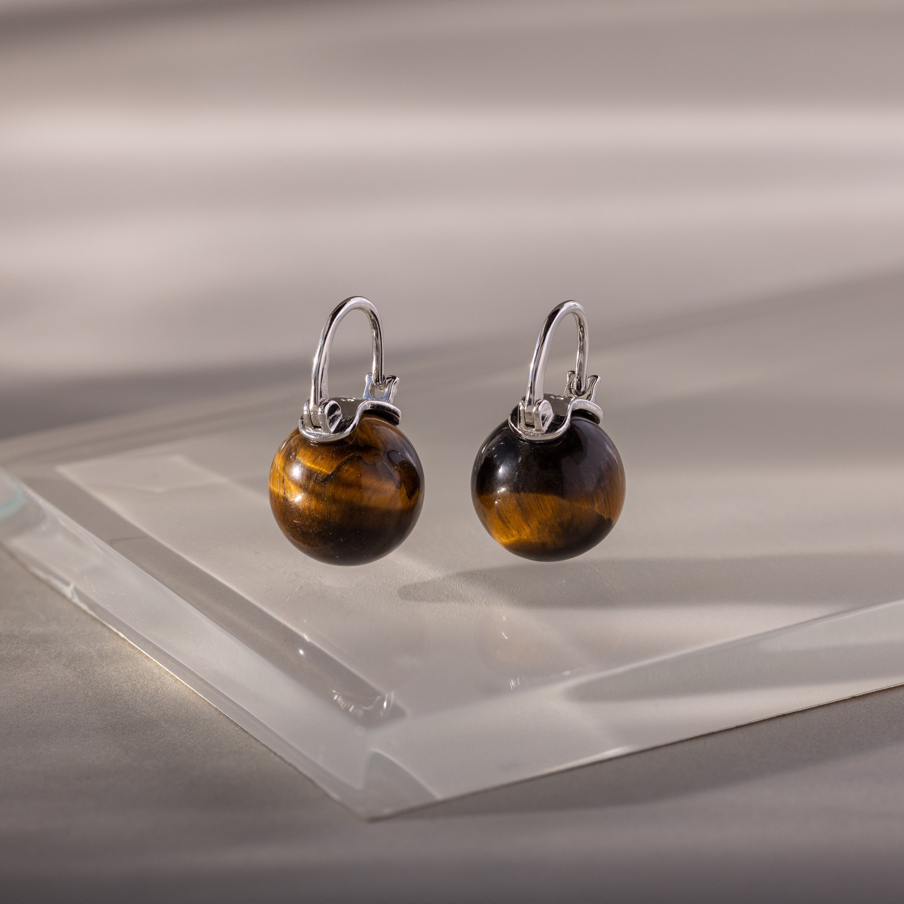 The Tiger's Eye Dome Drop Earrings feature round Tiger’s Eye gemstone beads on silver domes, showcased on a clear acrylic platform—a striking piece of natural stone jewelry.