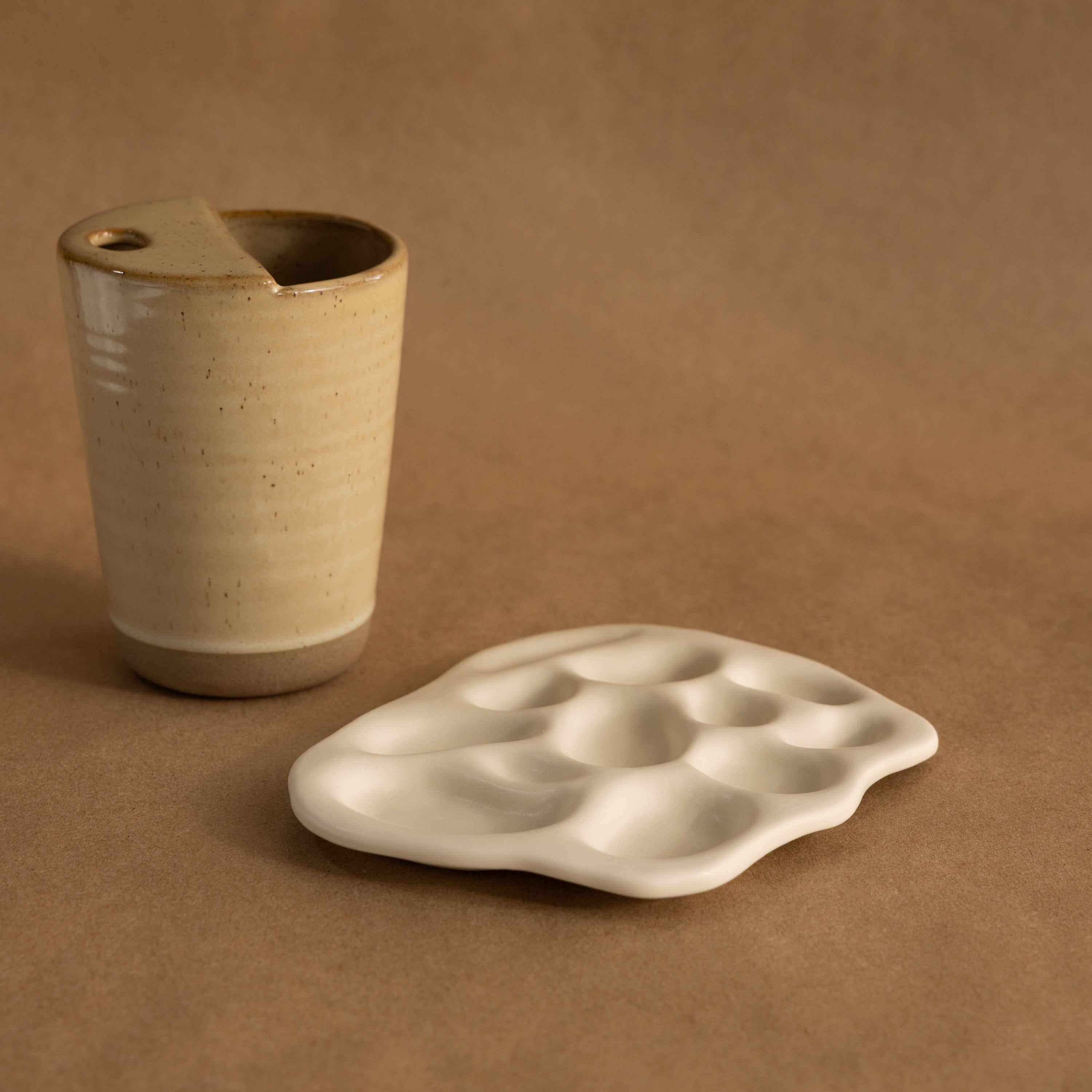 Beige ceramic cup and the Mini Organic Treasure Trove Jewelry Dish—a white, wavy ceramic piece perfect as a mini jewelry or keepsake holder—set on a brown background.