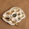 Gold and green jewelry—watch, rings, earrings, and necklace—elegantly arranged on the Mini Organic Treasure Trove Jewelry Dish with a wavy white design.