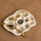 Gold and green jewelry—watch, rings, earrings, and necklace—elegantly arranged on the Mini Organic Treasure Trove Jewelry Dish with a wavy white design.