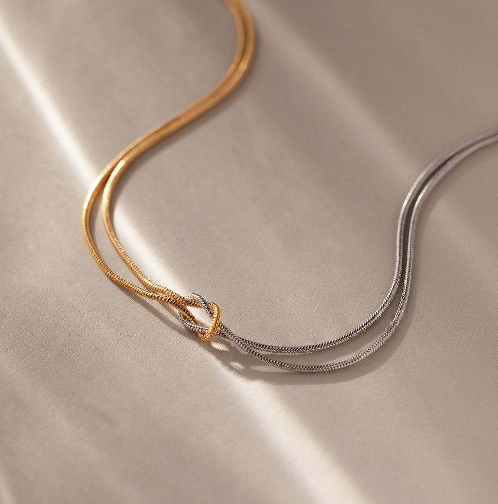 The Mixed Metal Knot Necklace features intertwined gold and silver chains, elegantly displayed on a soft beige surface under gentle lighting—a refined symbol of everlasting love.